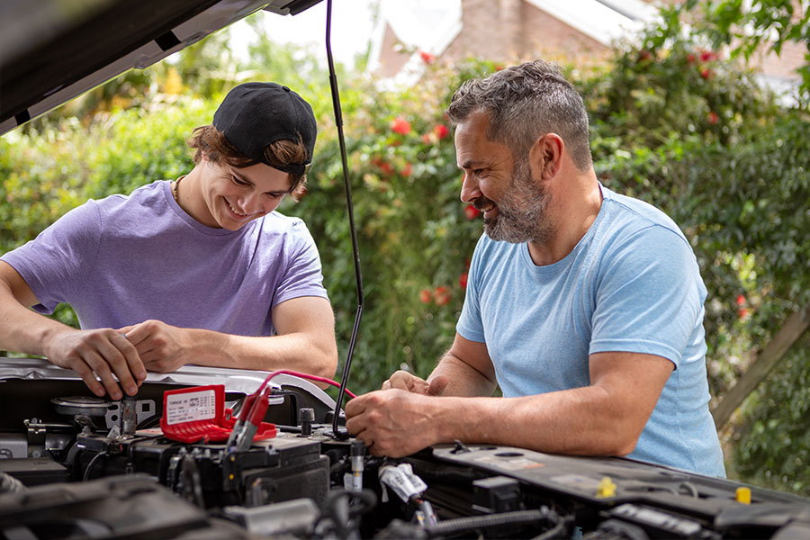 Father and son conducting auto maintenance under a car's hood.